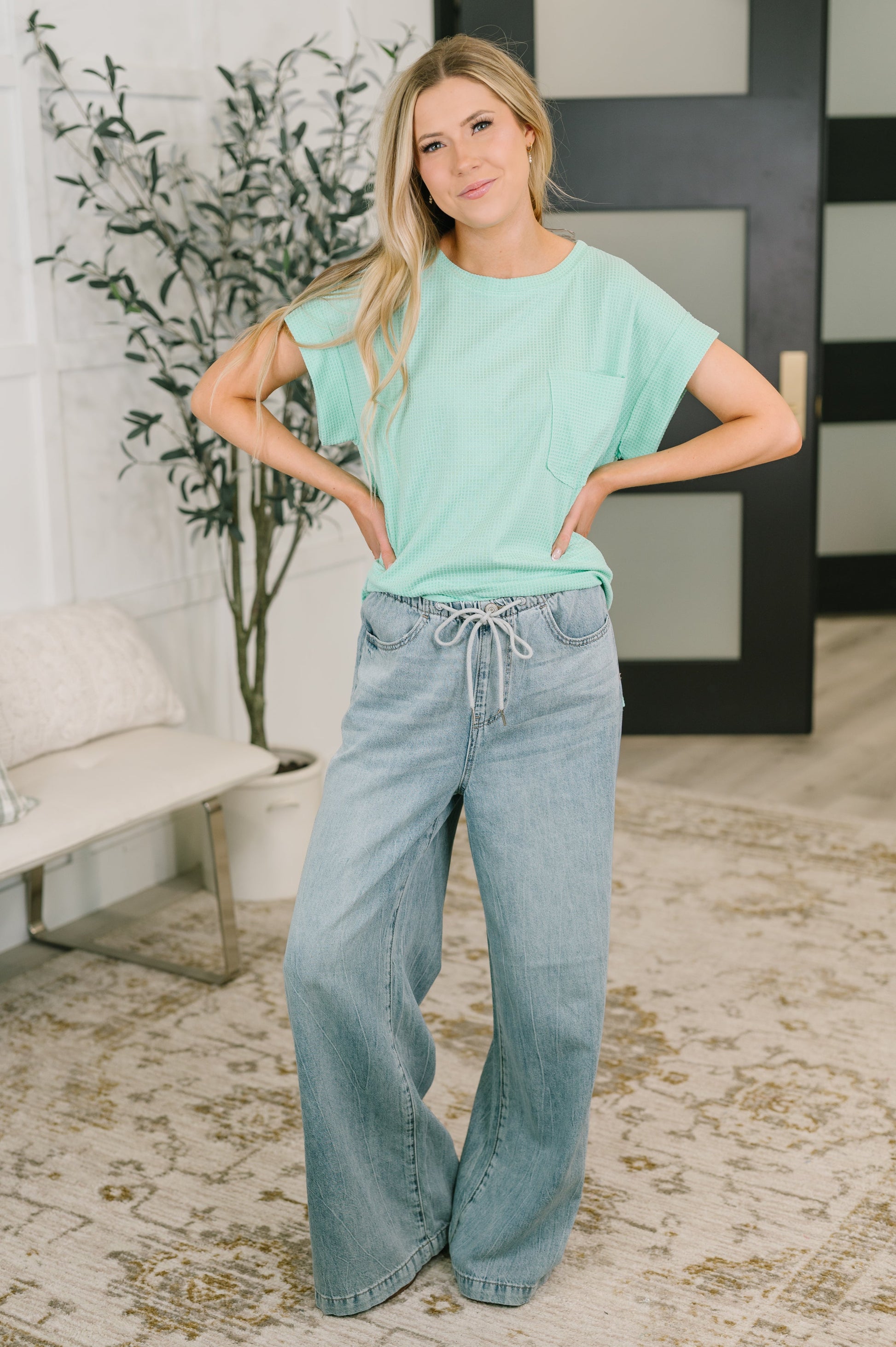 Woman wearing a light green top and blue jeans standing in a room with a plant and sofa.