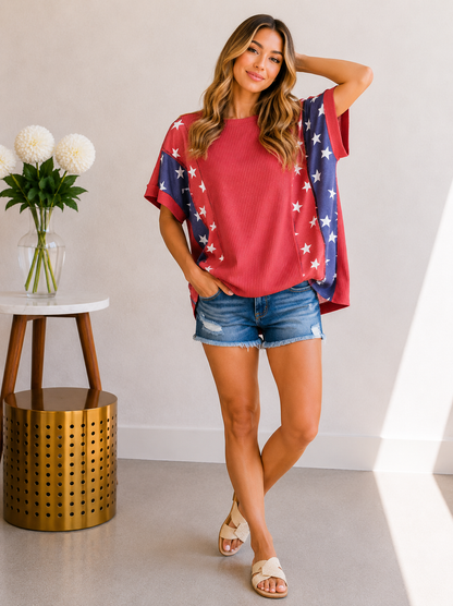 Woman wearing a red top with a blue and white pattern, denim shorts, and sandals in a room with a white wall and small table with flowers.