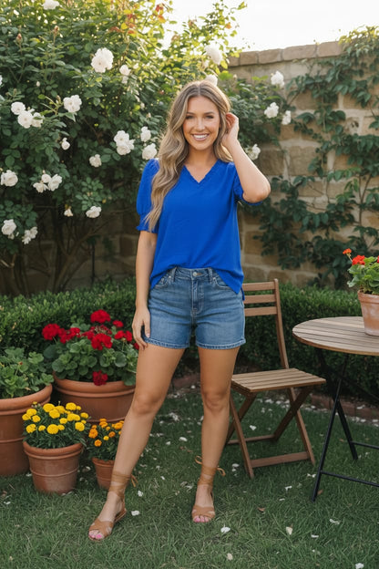 Woman in a blue shirt and denim shorts standing in a garden with flowers and a table in the background.
