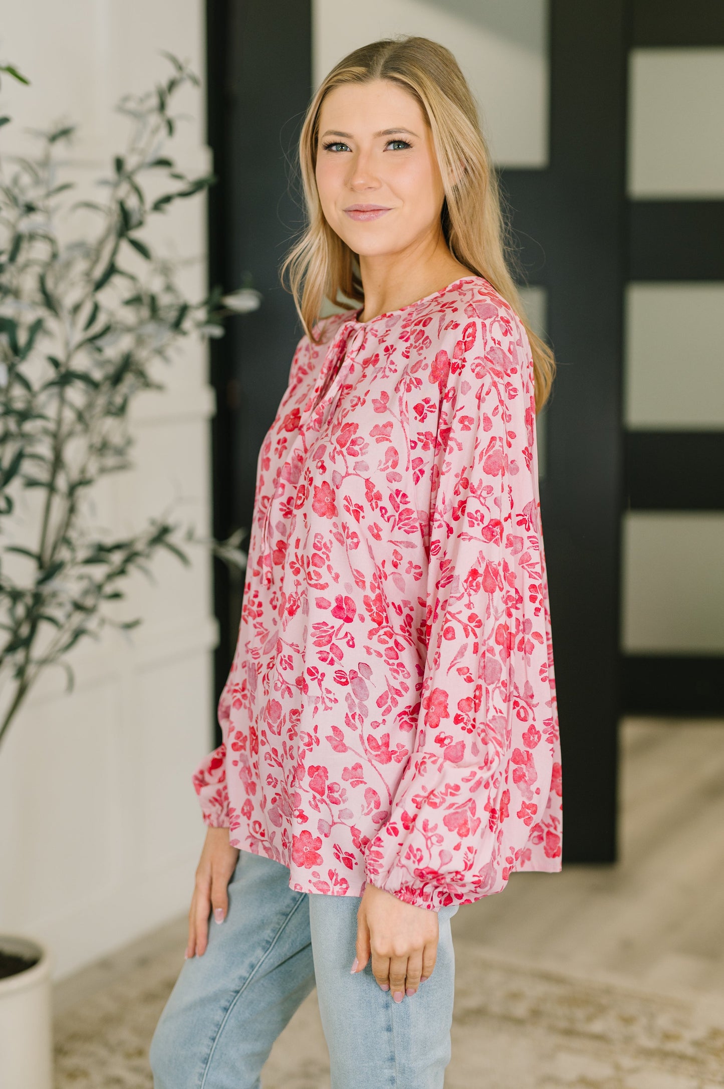 Woman wearing a pink floral blouse standing indoors with a plant and wall in the background