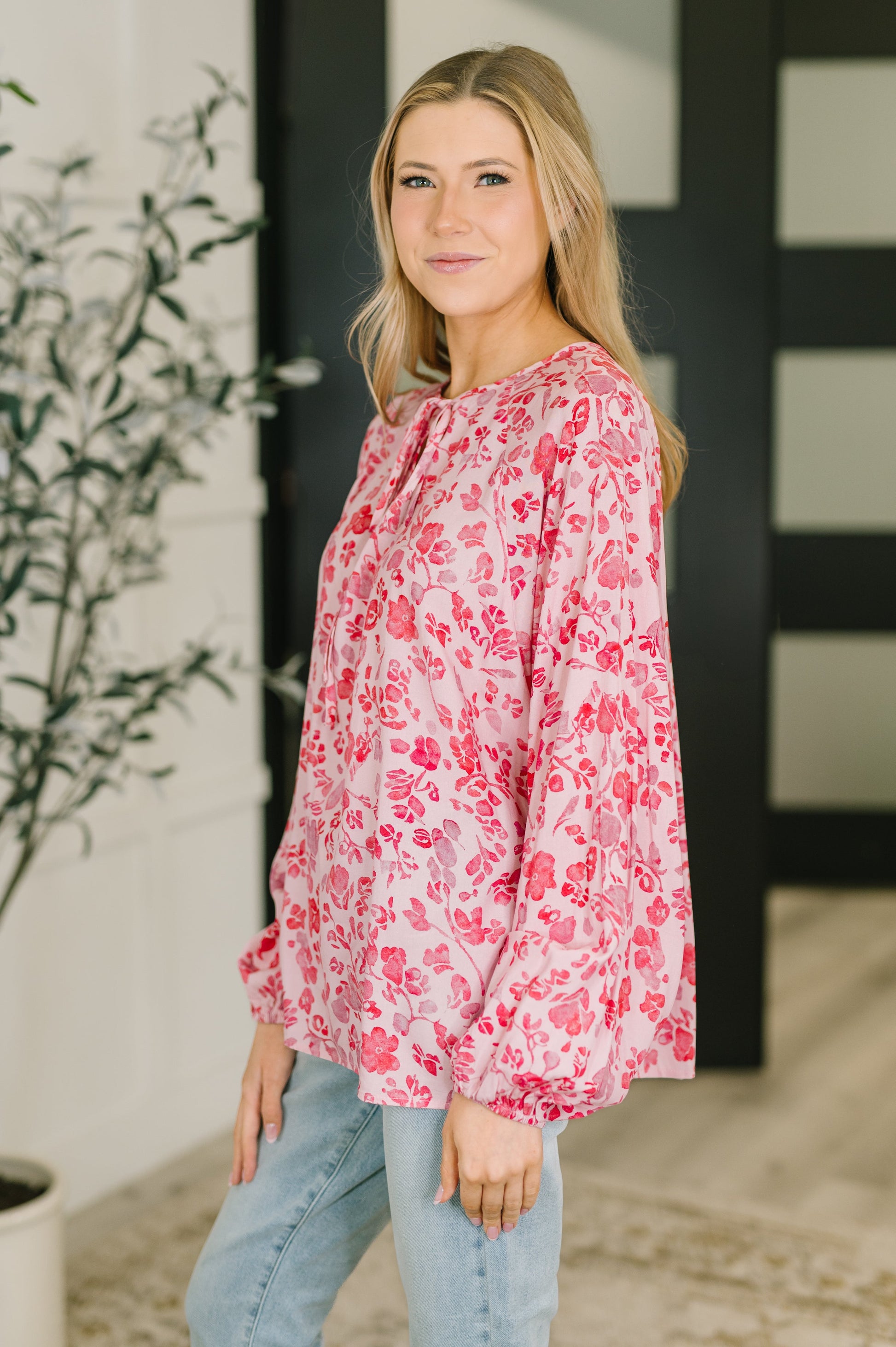 Woman wearing a pink floral blouse standing indoors with a plant and wall in the background