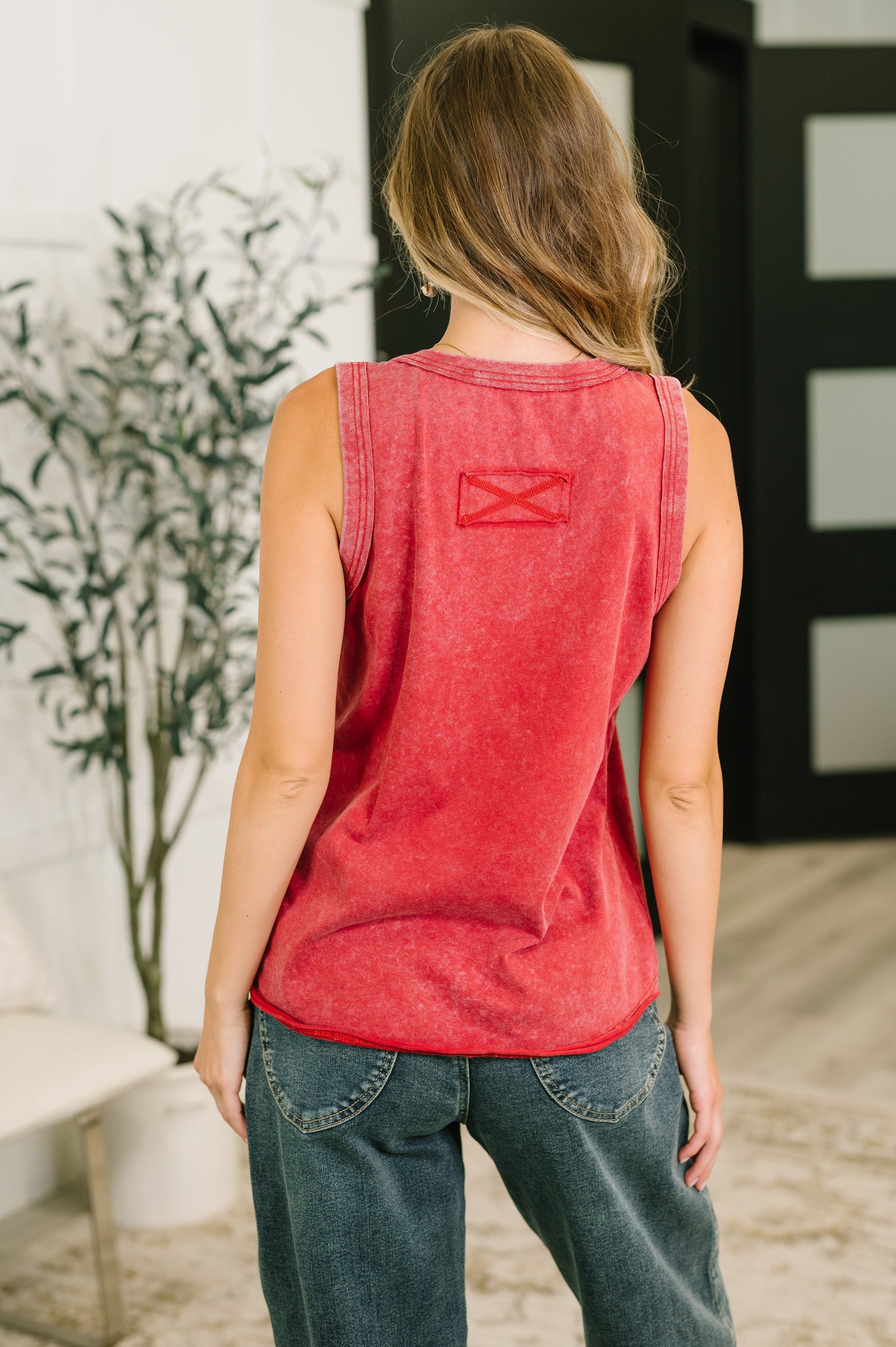 A woman viewed from behind wearing a red washed sleeveless tank top with a small red geometric logo on the upper back, paired with dark blue jeans in an indoor setting.