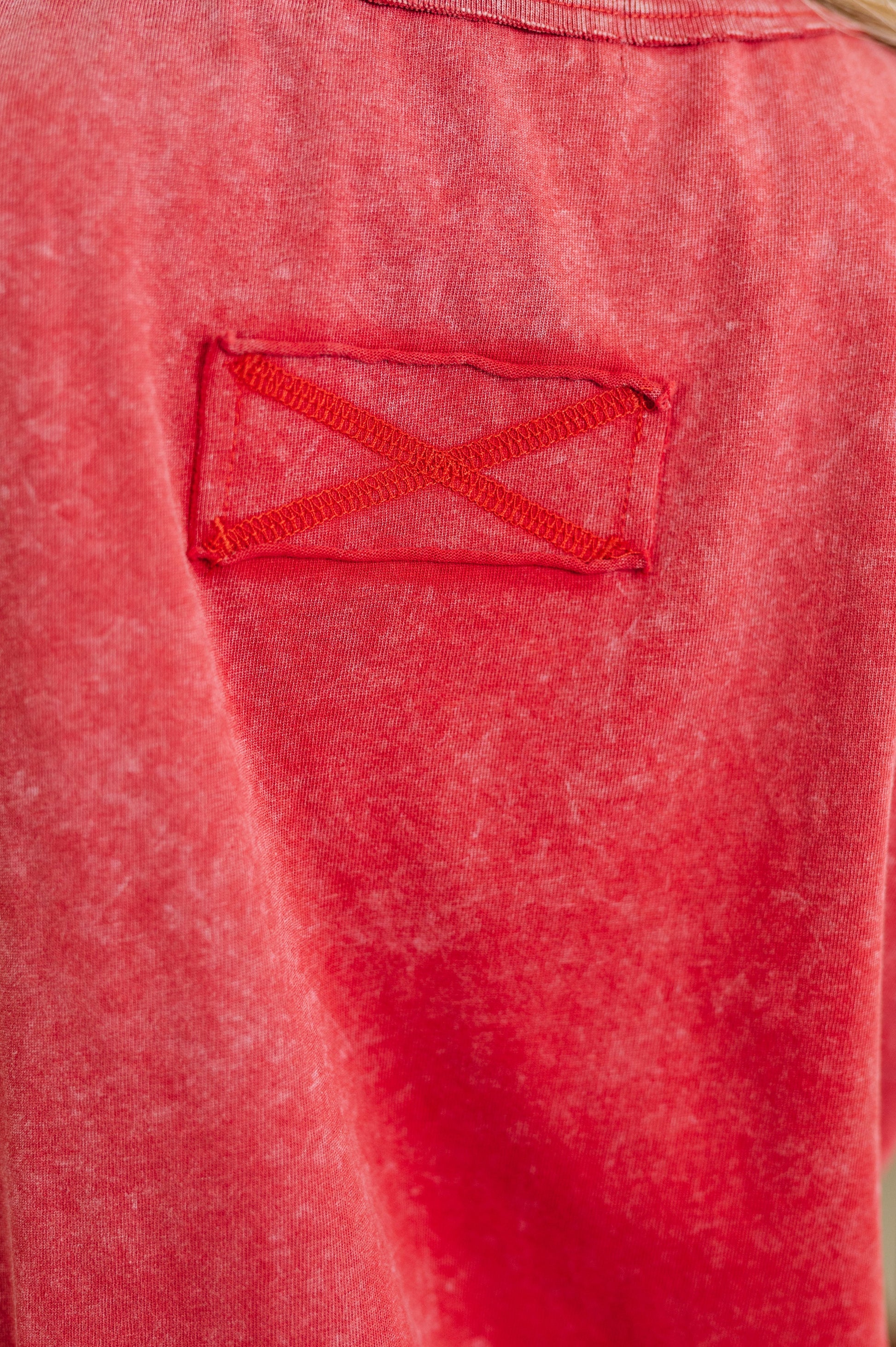 Close-up of a red textured fabric garment featuring a rectangular patch pocket with an embroidered red X-shaped detail.