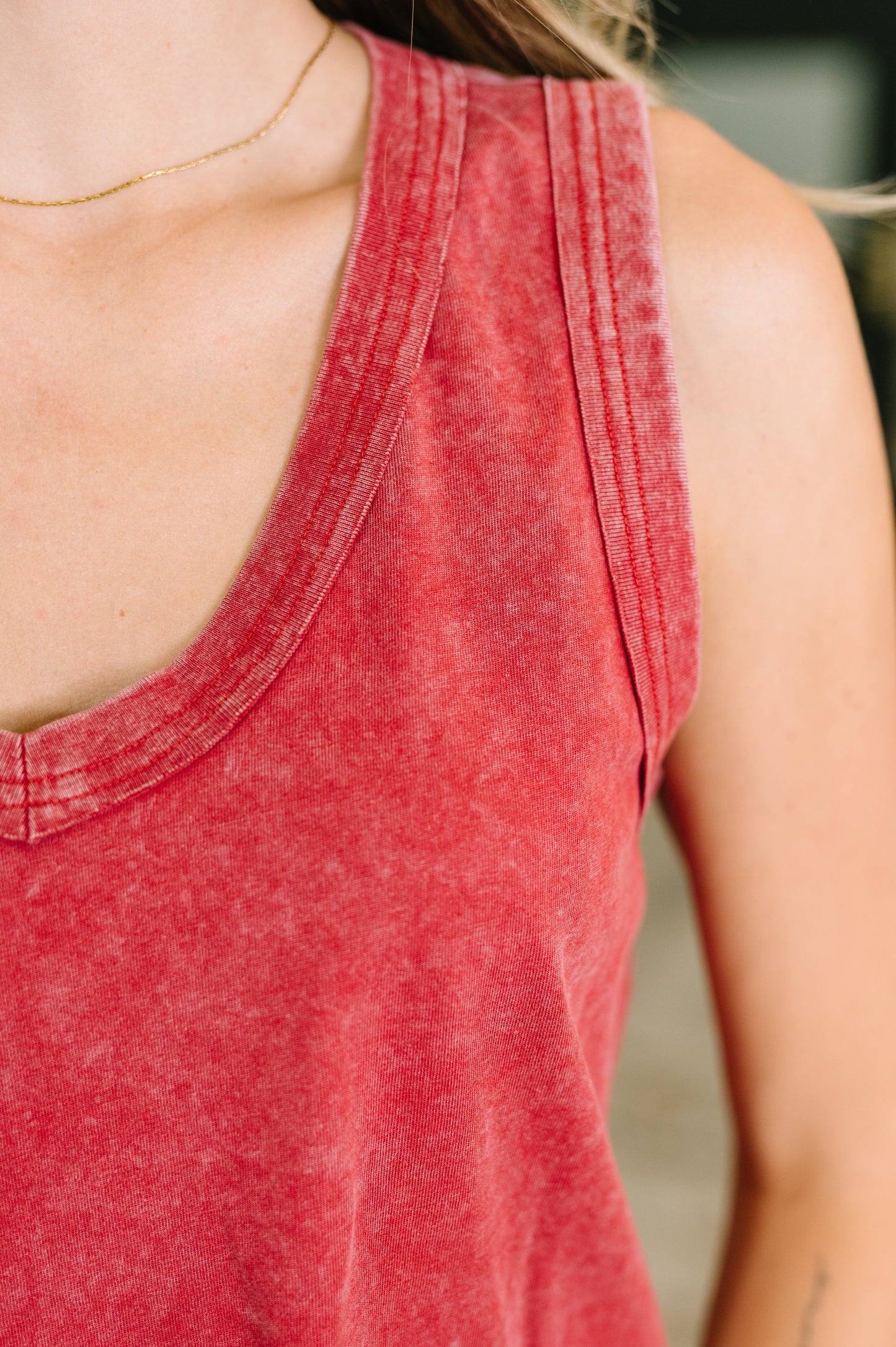 Close-up of a person wearing a washed red racerback tank top, showing the shoulder, chest, and neckline against a softly blurred background.