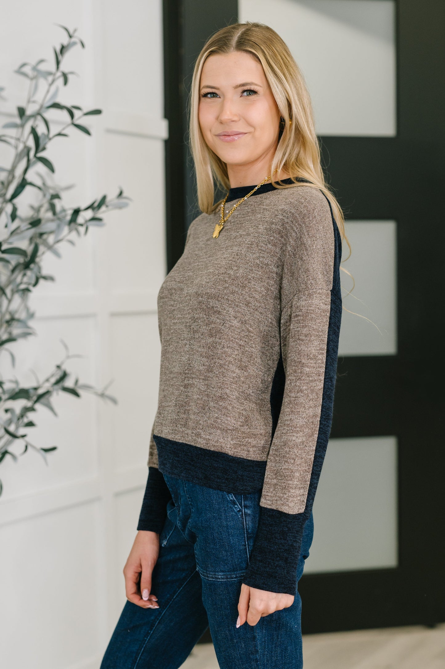 Woman wearing a two-tone sweater and jeans standing indoors with a plant in the background