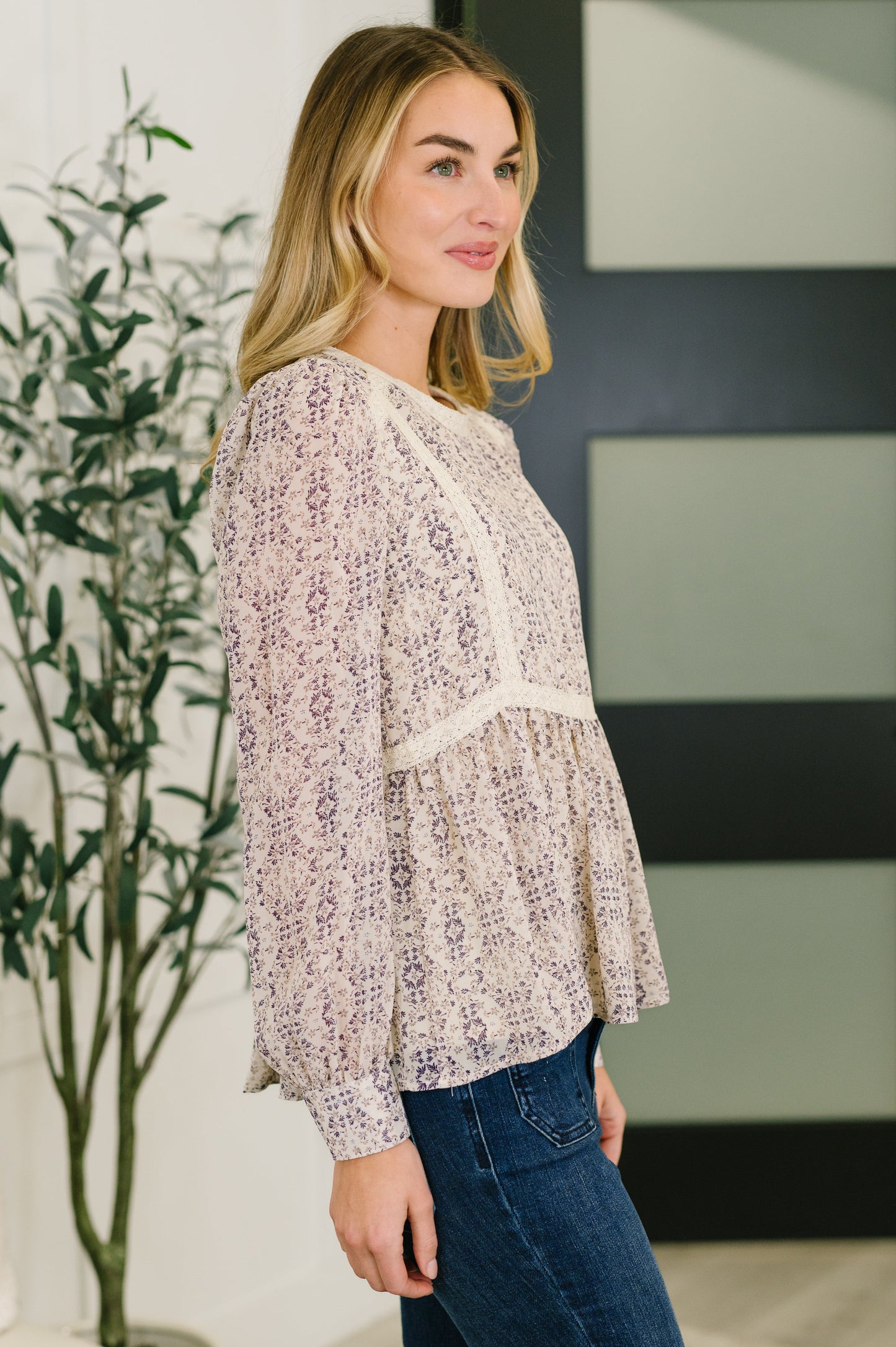 Woman wearing a floral blouse and jeans standing indoors with a plant in the background