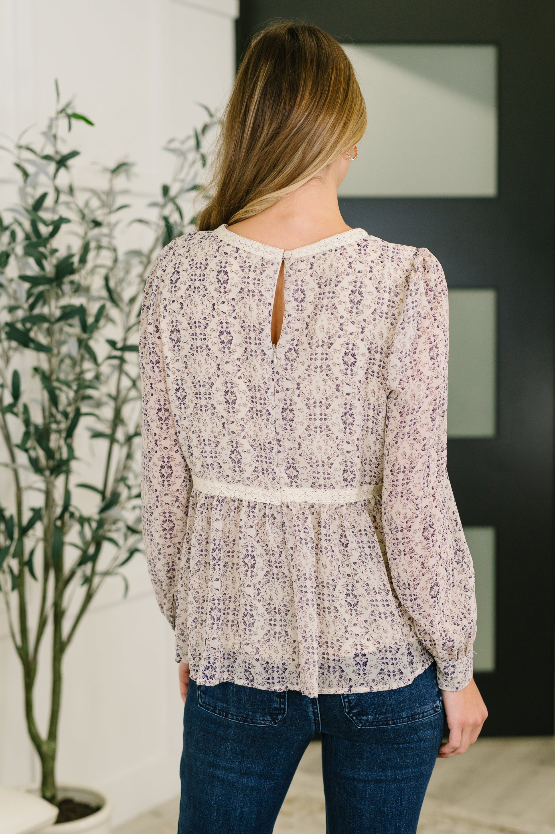 Woman wearing a patterned blouse and jeans standing indoors with a plant in the background