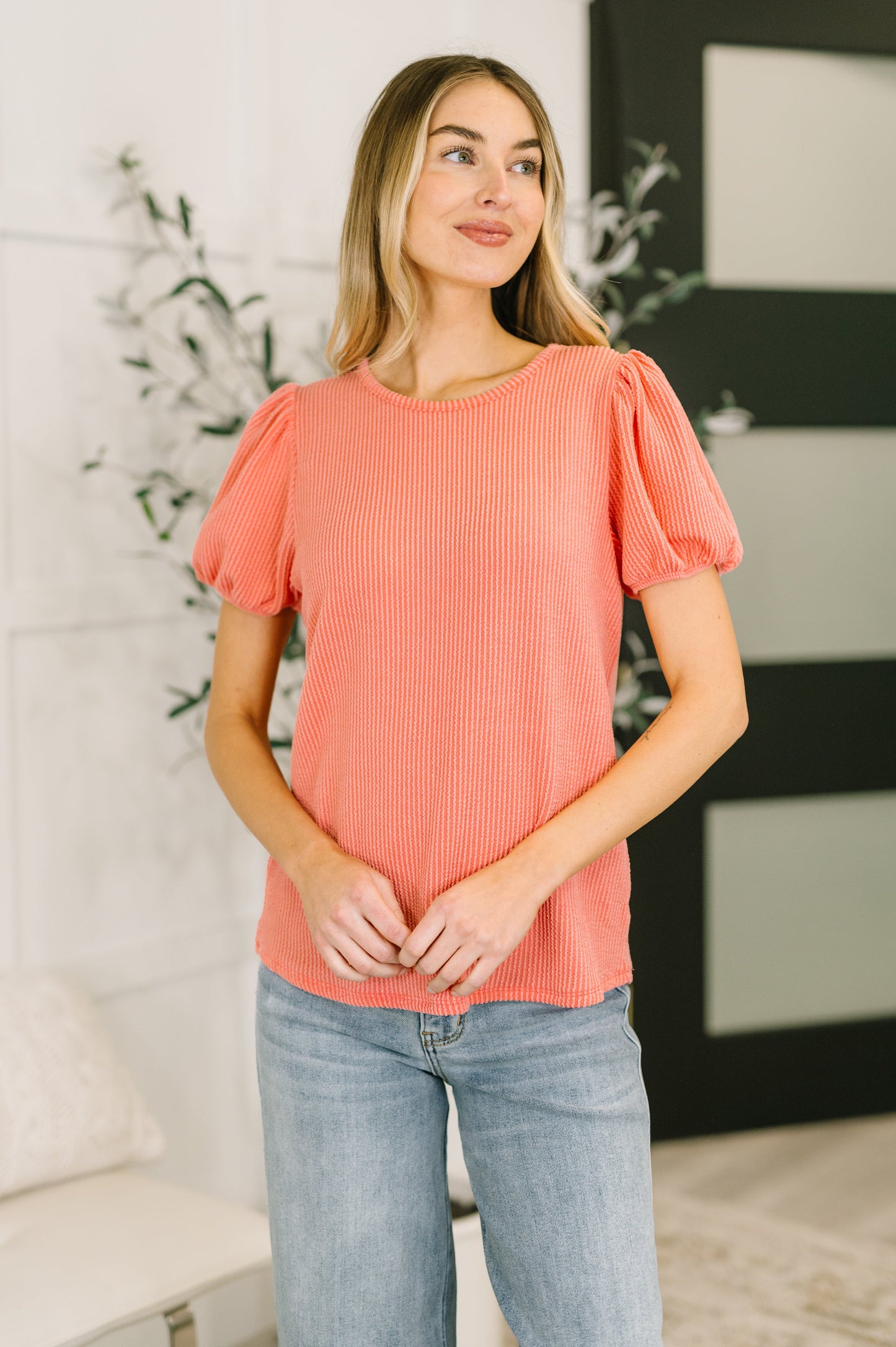 Woman wearing a coral top and blue jeans standing indoors.