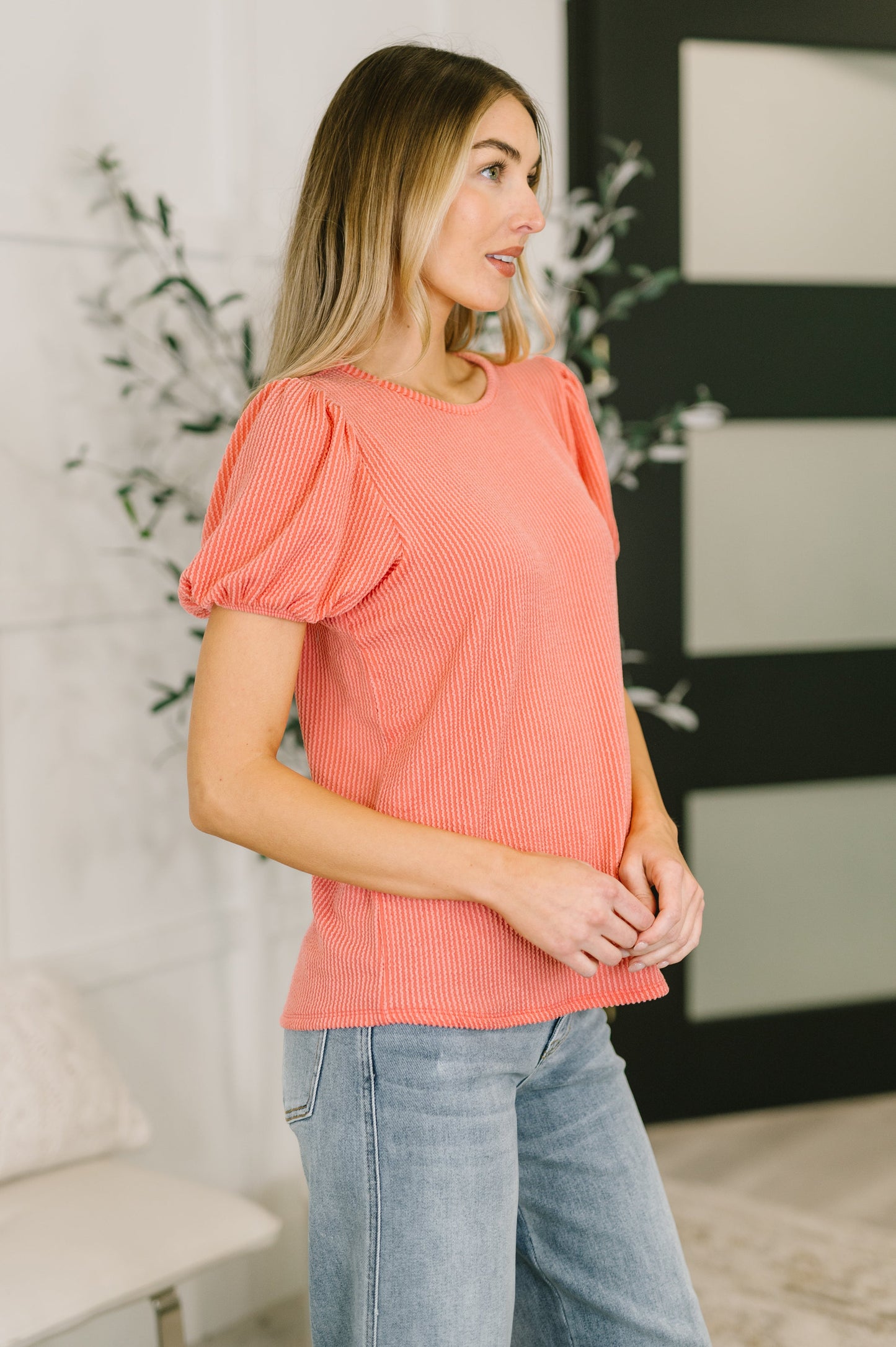 Woman wearing a coral top and Judy Blue jeans standing indoors with decorative elements in the background