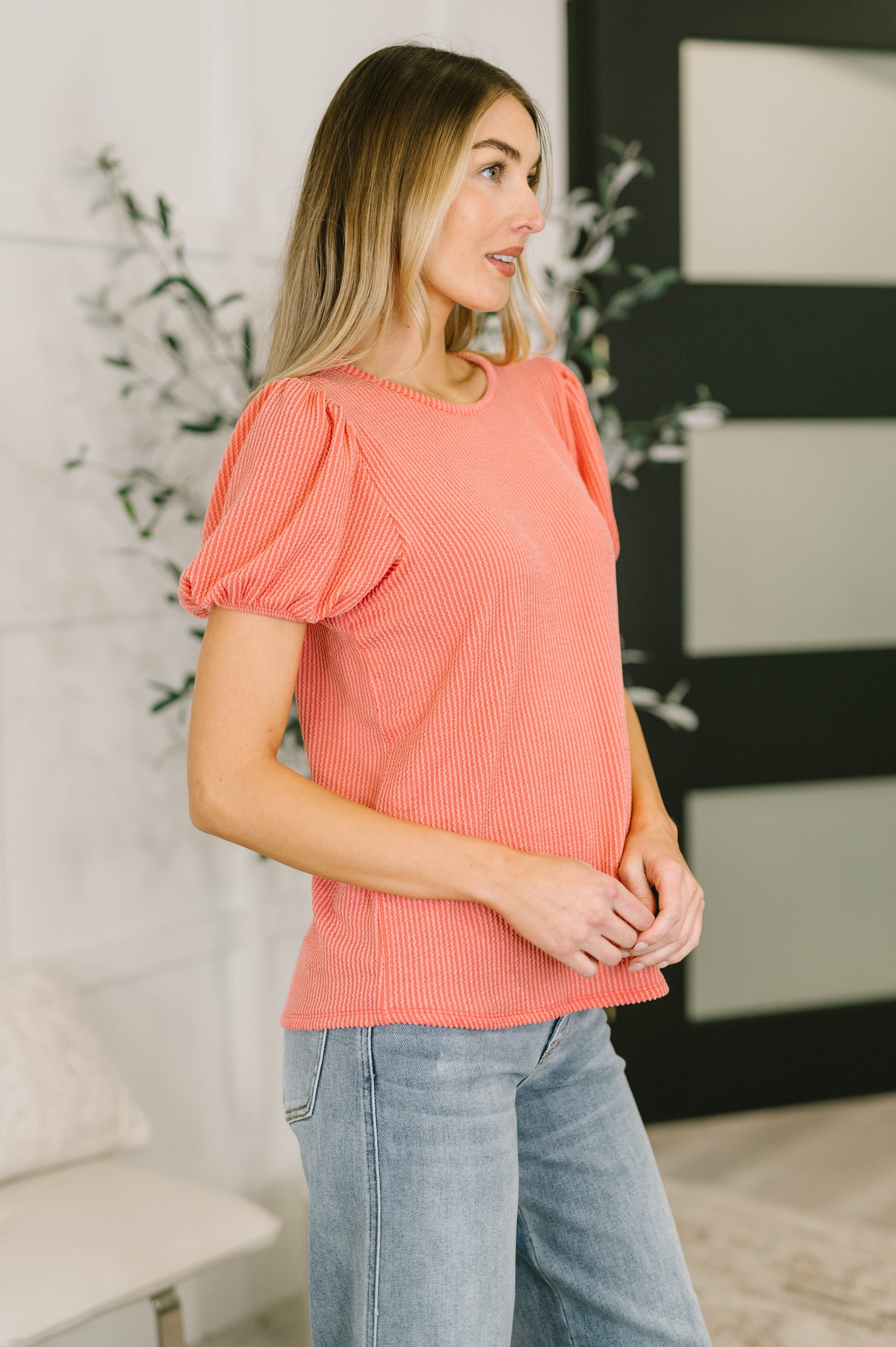 Woman wearing a coral top and Judy Blue jeans standing indoors with decorative elements in the background