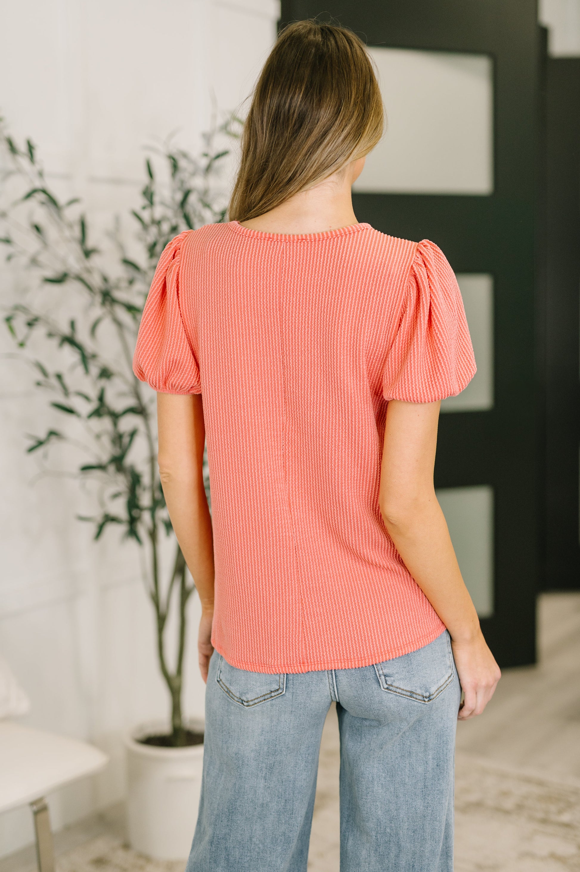 Woman wearing a coral top and light wash Judy Blue jeans standing indoors with a plant in the background