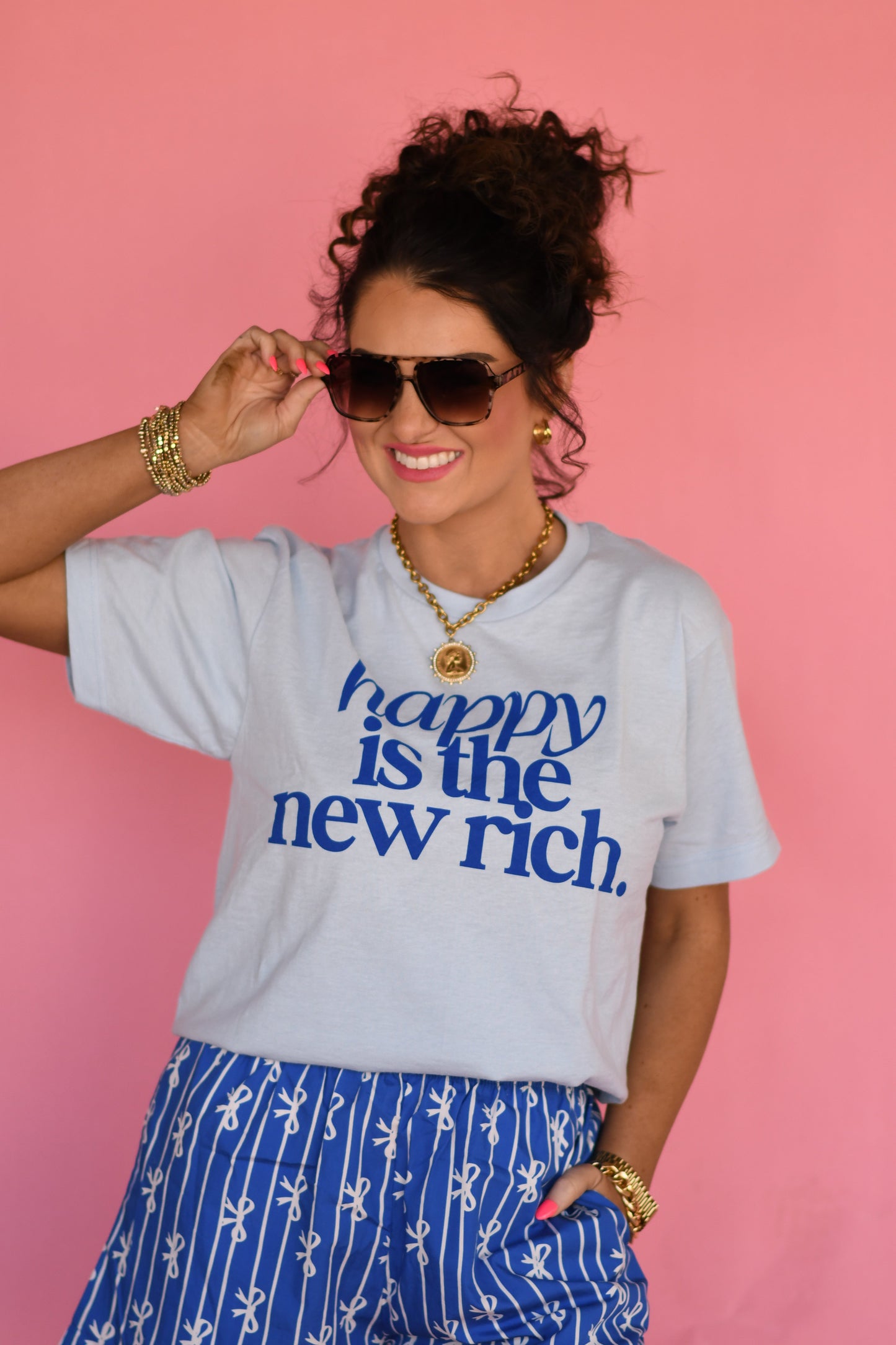 Woman wearing a light blue t-shirt with 'happy is the new rich' text, sunglasses, and gold jewelry against a pink background.
