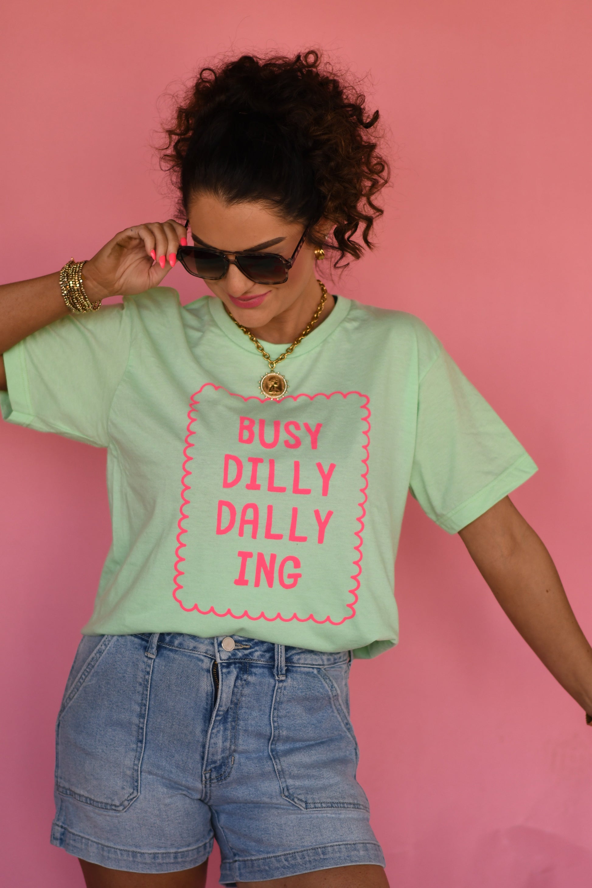 Woman wearing a light green t-shirt with 'BUSY DILLY DALLYING' text on a pink background