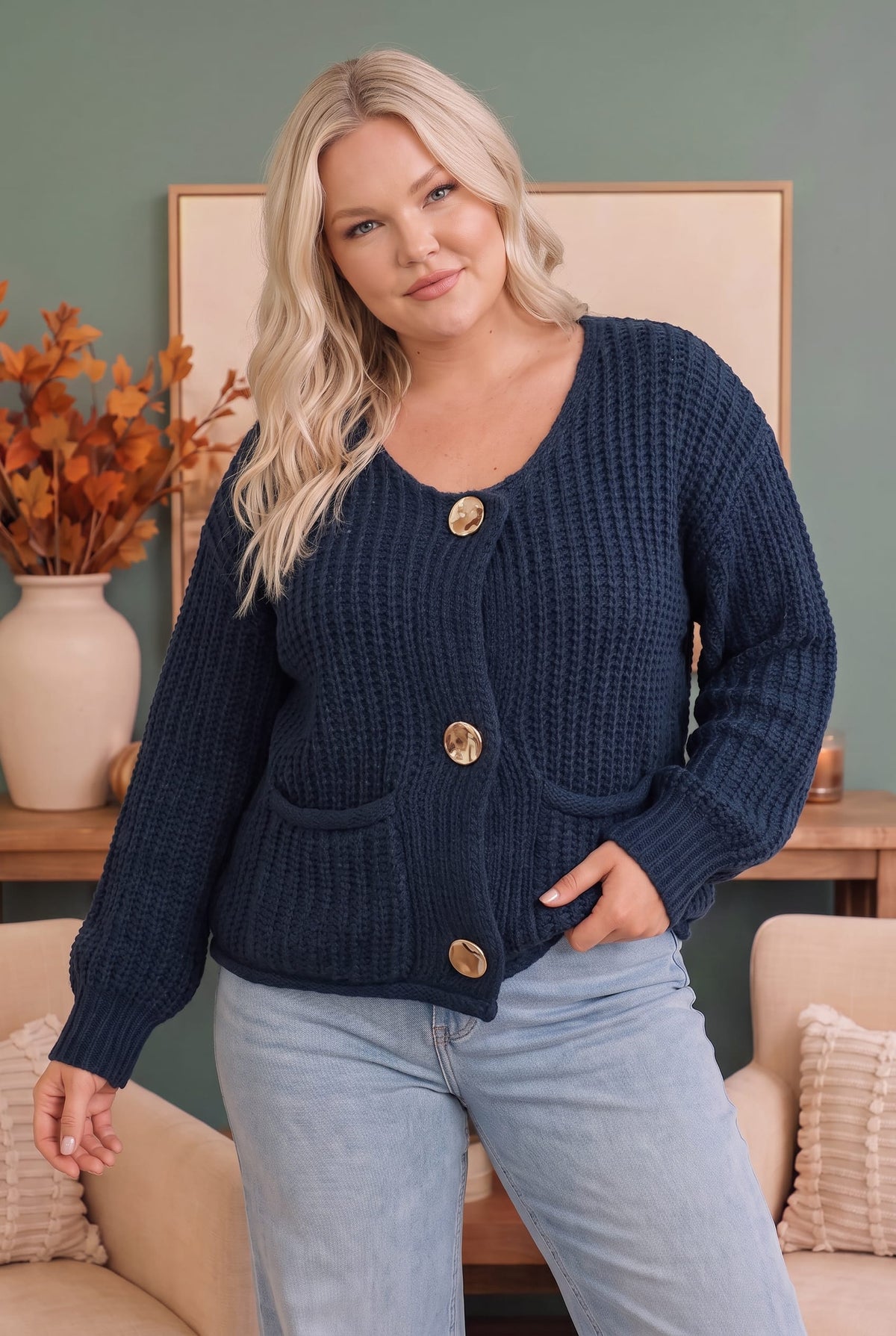 A woman wearing a navy blue chunky cable-knit cardigan with gold buttons and front pockets, paired with light blue jeans, standing in a cozy living room setting.