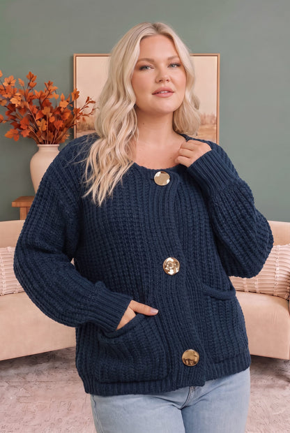 A woman wearing a chunky navy blue cable-knit cardigan sweater with large gold buttons, paired with light blue jeans, standing in a cozy living room setting.
