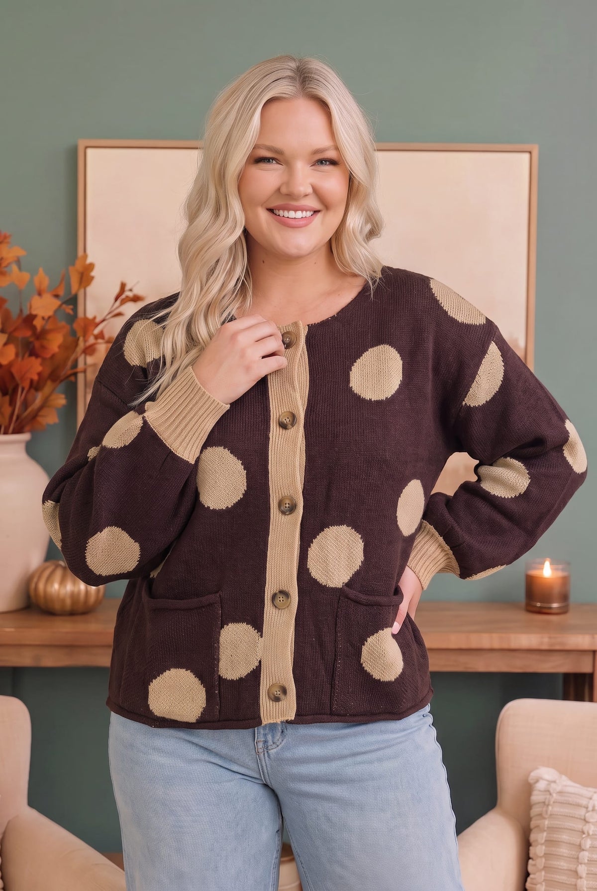 A woman wearing a dark brown cardigan with large beige polka dots and beige ribbed trim, paired with light blue jeans, standing in a cozy indoor setting.