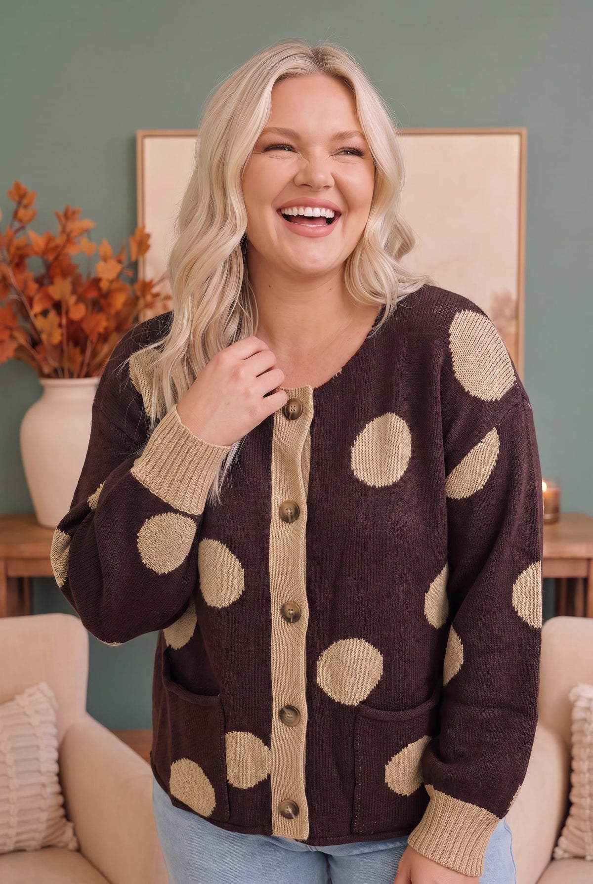 A woman smiling while wearing a dark brown cardigan sweater with beige polka dots and ribbed trim, standing in a cozy living room setting.