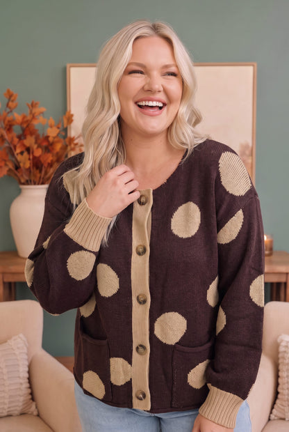 A woman smiling while wearing a dark brown cardigan sweater with beige polka dots and ribbed trim, standing in a cozy living room setting.