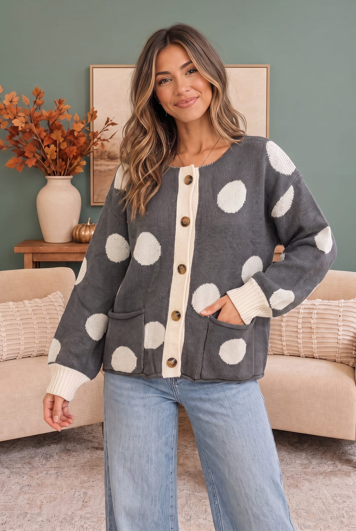 A woman wearing a gray cardigan with large white polka dots and cream ribbed trim, paired with light blue jeans, standing in a living room setting.