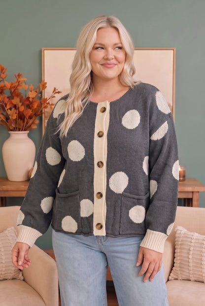 A woman wearing a gray cardigan with large cream polka dots and a cream button placket, paired with light blue jeans, standing in a cozy living room setting.