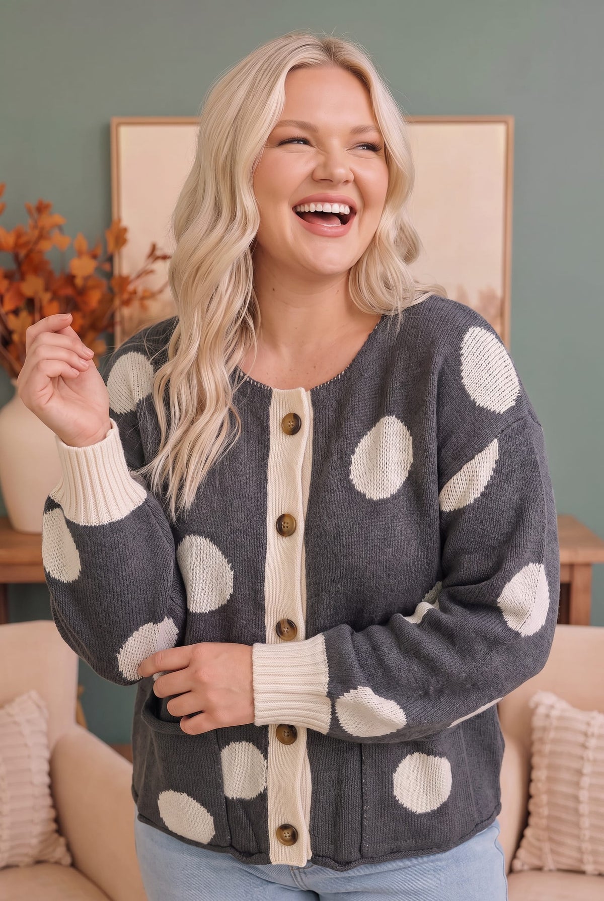 A woman wearing a dark gray knit cardigan with large cream polka dots and a cream button placket, standing in a cozy indoor setting.
