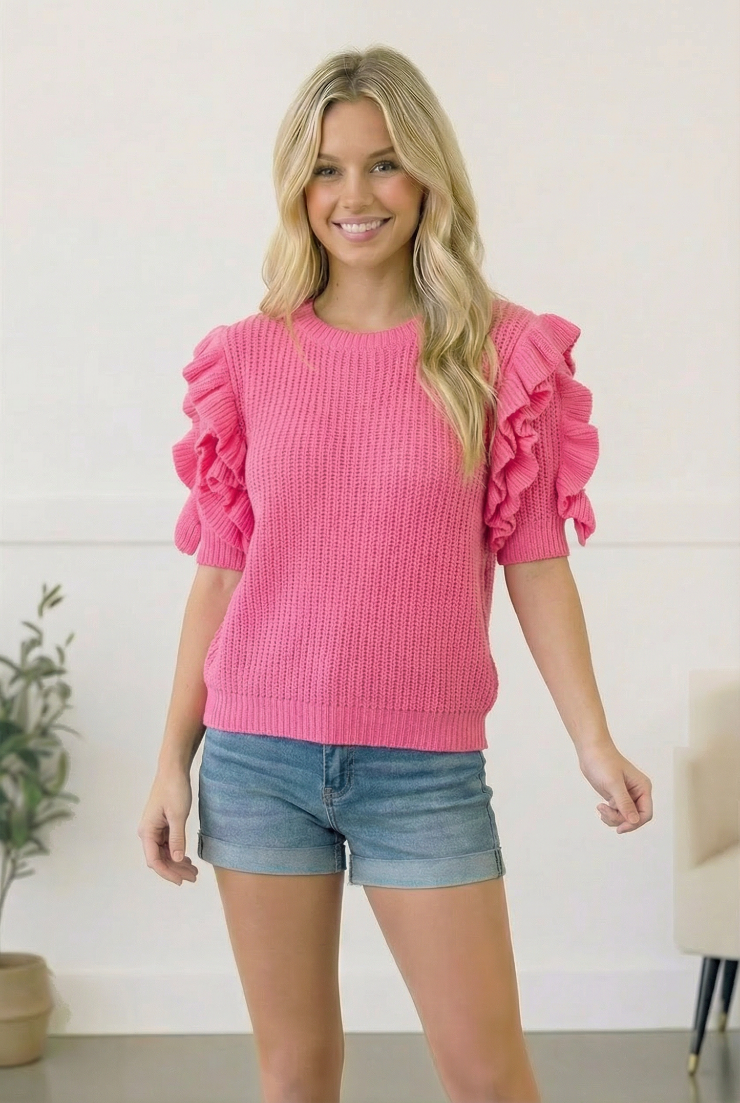 Woman wearing a pink sweater with ruffled sleeves and denim shorts indoors.