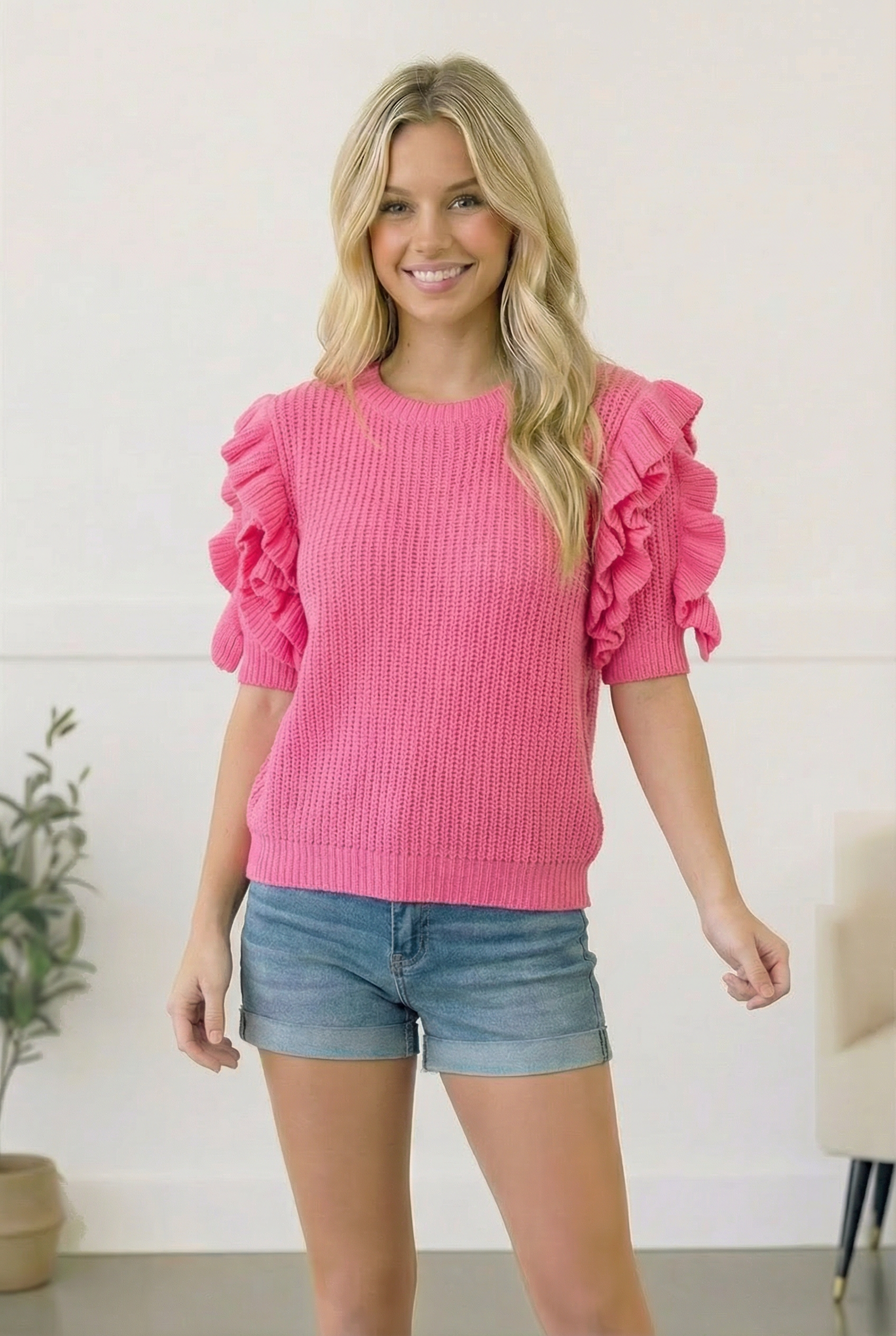 Woman wearing a pink sweater with ruffled sleeves and denim shorts indoors.