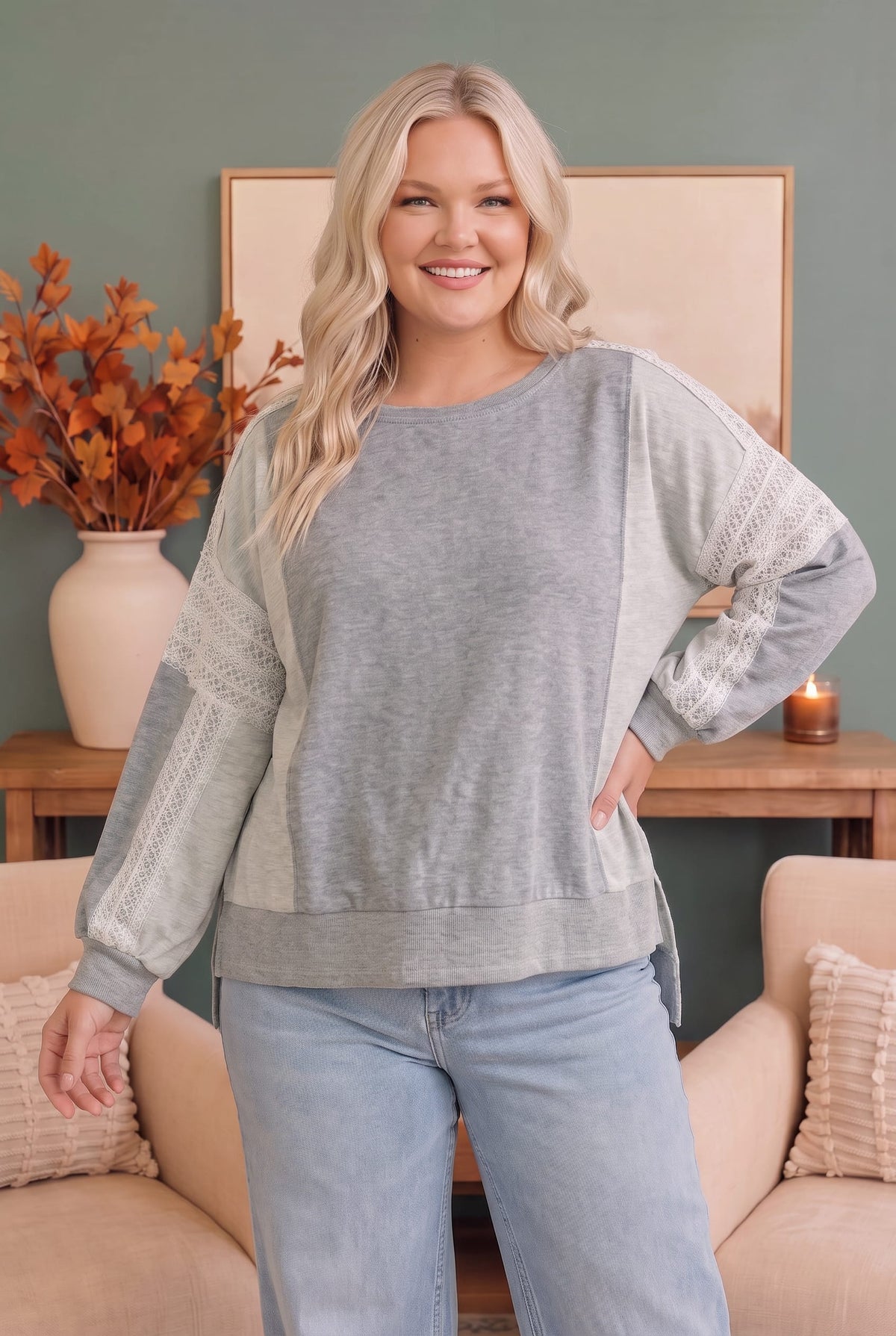 A woman modeling a heather gray long-sleeve top with textured lace-like panels on the sleeves, paired with light blue jeans in a cozy living room setting.