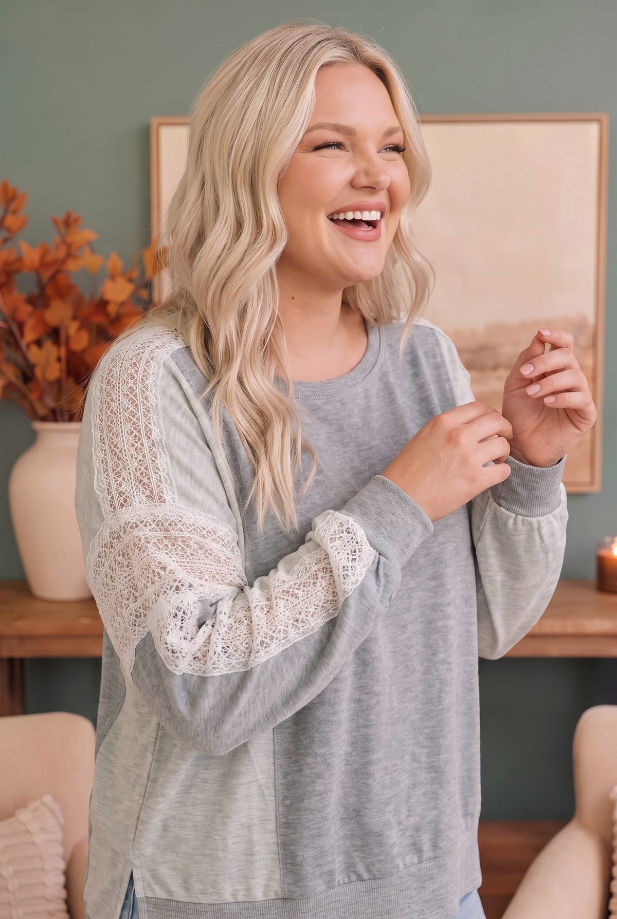 A woman with blonde hair smiling and laughing while wearing a heather gray long-sleeve top with white lace sleeves, set in a cozy indoor room with a framed picture and autumn decor.