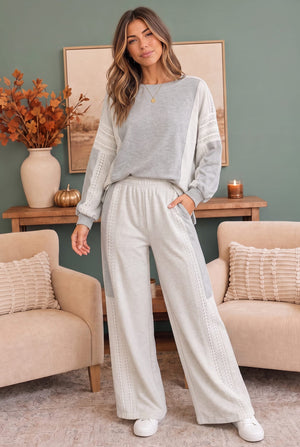A woman modeling a two-piece loungewear set consisting of a light gray long-sleeve top with textured side panels and matching off-white wide-leg pants with lace-up details, set in a cozy living room environment.