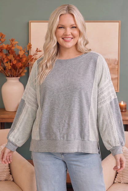 A woman modeling a light gray long-sleeve sweatshirt with textured knit lace sleeves, paired with light blue jeans in a cozy indoor setting.