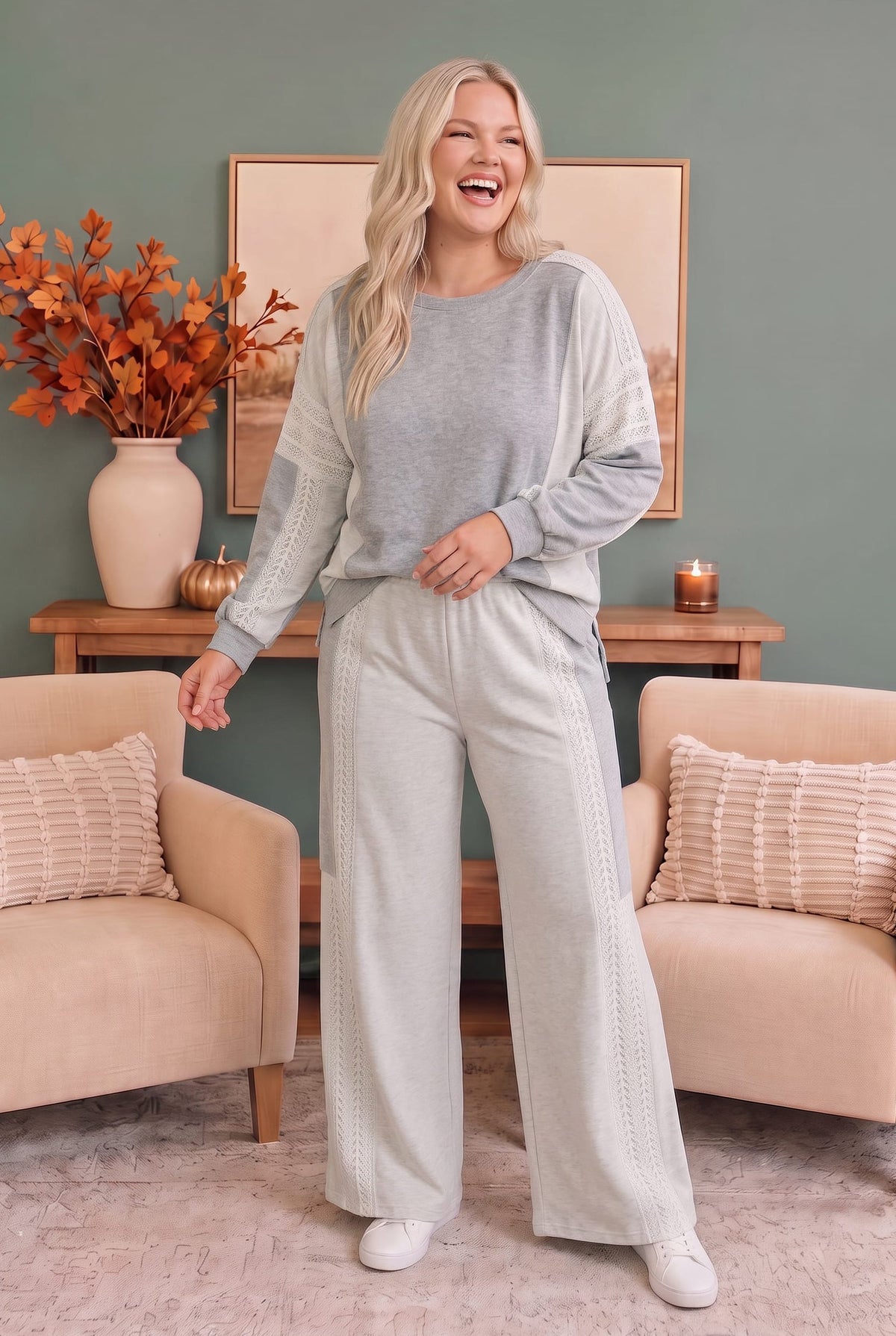 A woman modeling a light gray and white knit lounge set consisting of a long-sleeve top and matching wide-leg pants, standing in a cozy living room setting.