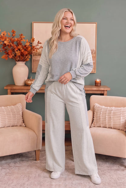 A woman modeling a light gray and white knit lounge set consisting of a long-sleeve top and matching wide-leg pants, standing in a cozy living room setting.