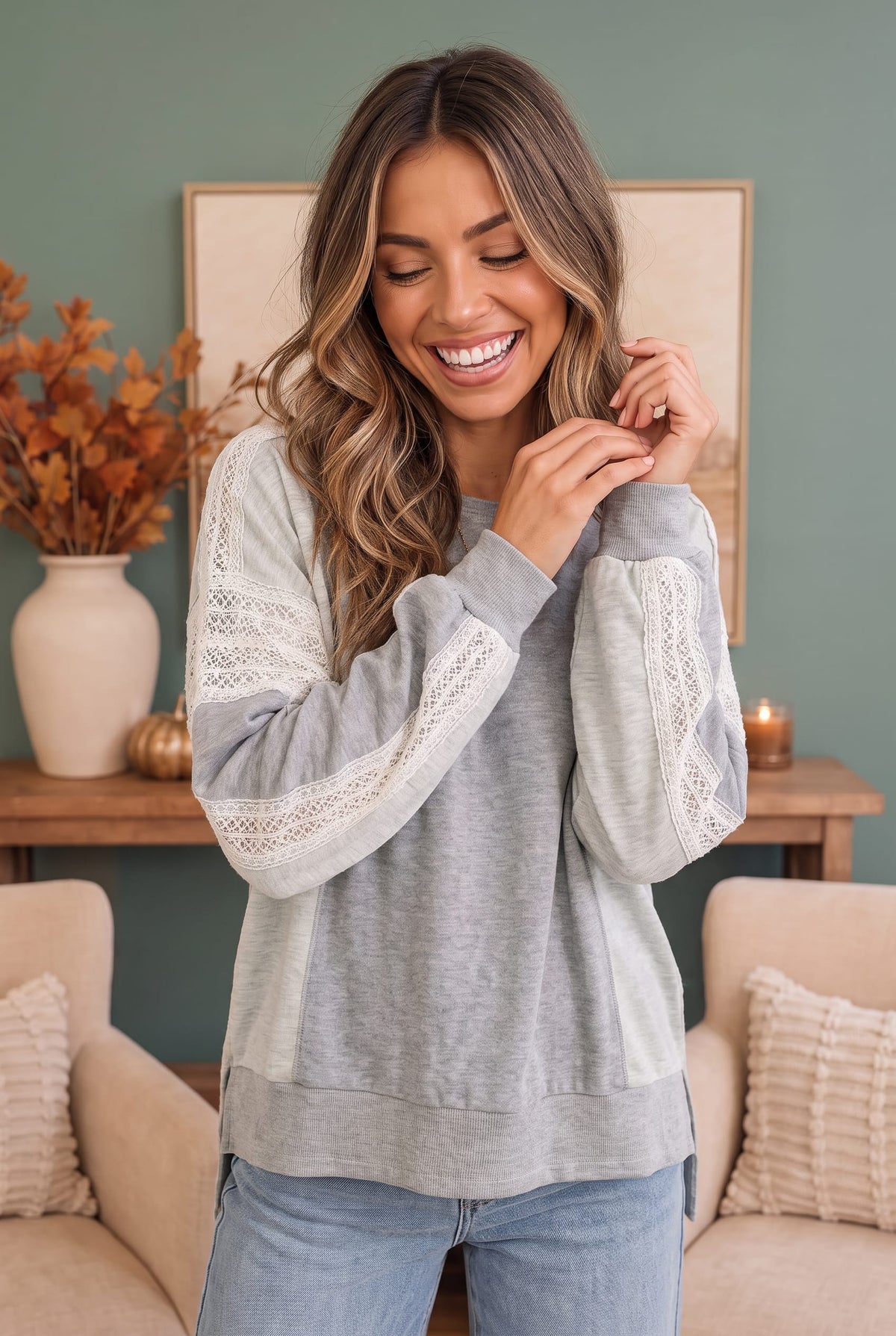 A woman wearing a light gray and white color-blocked sweatshirt with lace sleeve details, paired with light blue jeans, standing in a cozy living room setting.