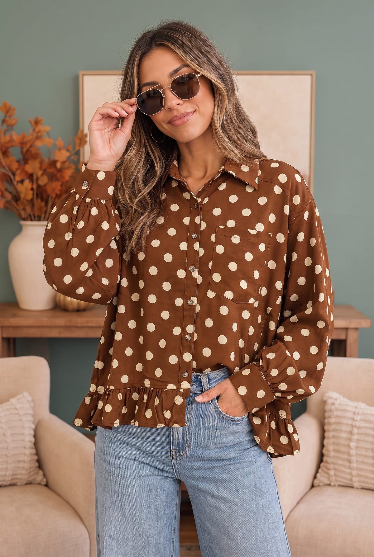 A woman wearing a brown long-sleeve button-up blouse with beige polka dots, paired with light blue jeans, standing in a cozy living room setting.