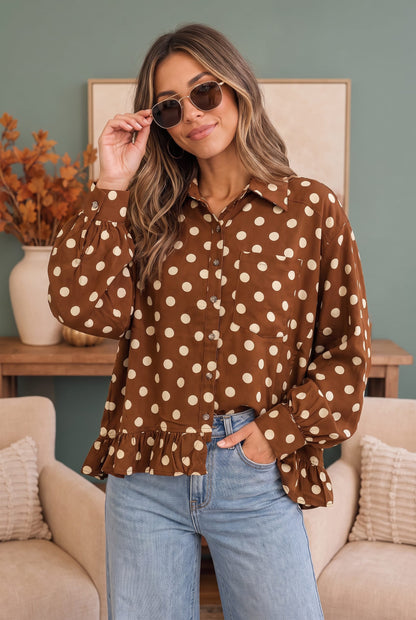 A woman wearing a brown long-sleeve button-up blouse with beige polka dots, paired with light blue jeans, standing in a cozy living room setting.