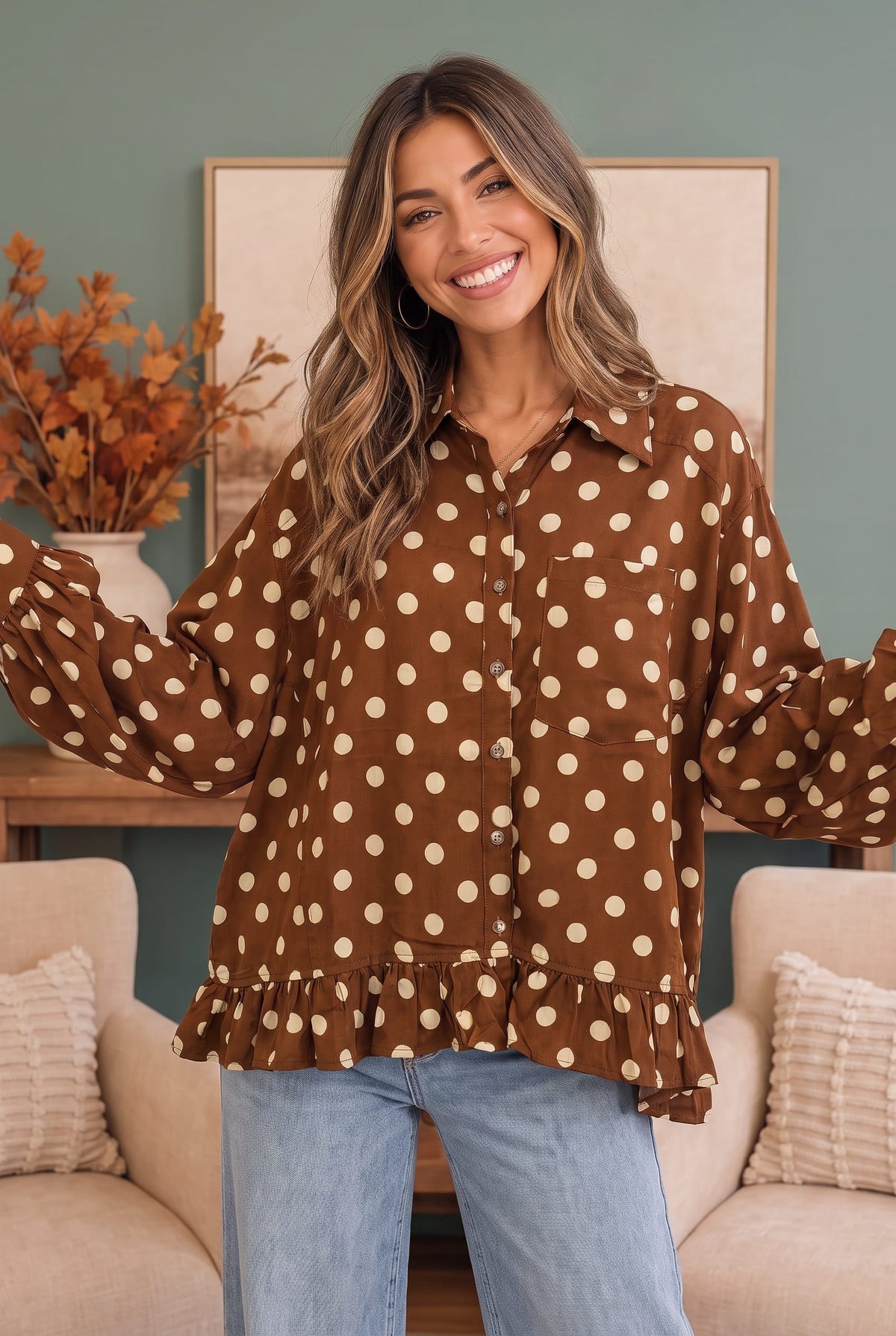 A woman wearing a brown long-sleeve button-up blouse with beige polka dots and a ruffled hem, paired with light blue jeans, standing in a living room setting.