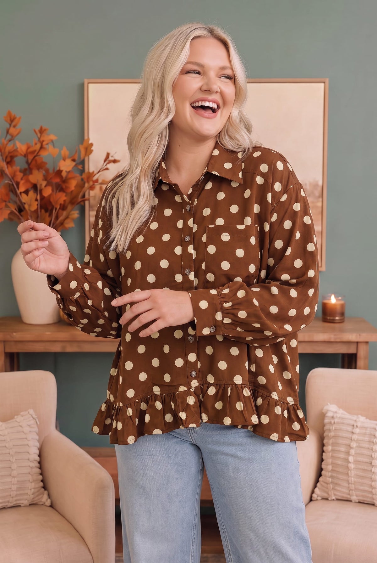 A woman modeling a brown long-sleeve button-up blouse with beige polka dots, paired with light blue jeans, standing in a cozy room setting with a teal wall, framed art, and beige armchairs.