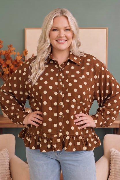 A woman modeling a brown long-sleeve button-up blouse with beige polka dots and a ruffled hem, paired with light blue jeans, standing in a styled indoor setting.