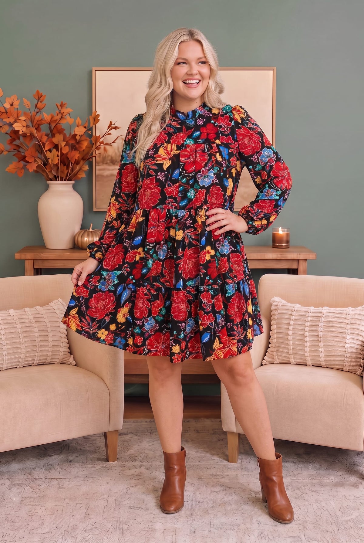 A woman modeling a long-sleeve high-neck mini dress with a vibrant red, blue, and yellow floral print on a dark background, standing in a cozy living room setting.