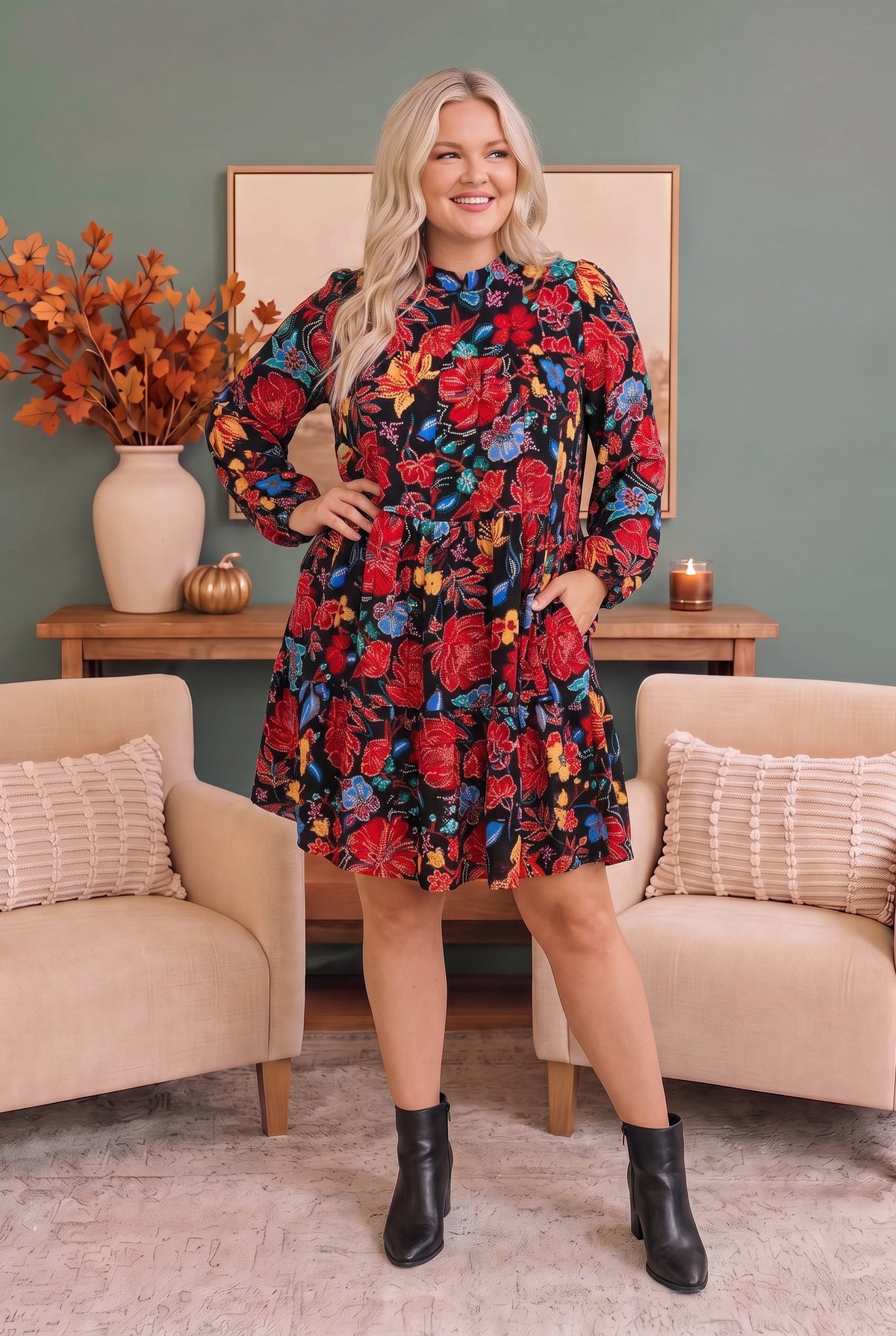 A woman modeling a long-sleeve high-neck dress with a vibrant red, blue, and yellow floral pattern, standing in a styled living room setting.