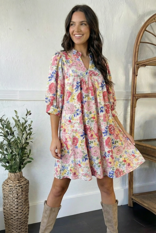Woman wearing a floral dress standing in a room with a plant and wooden shelf.