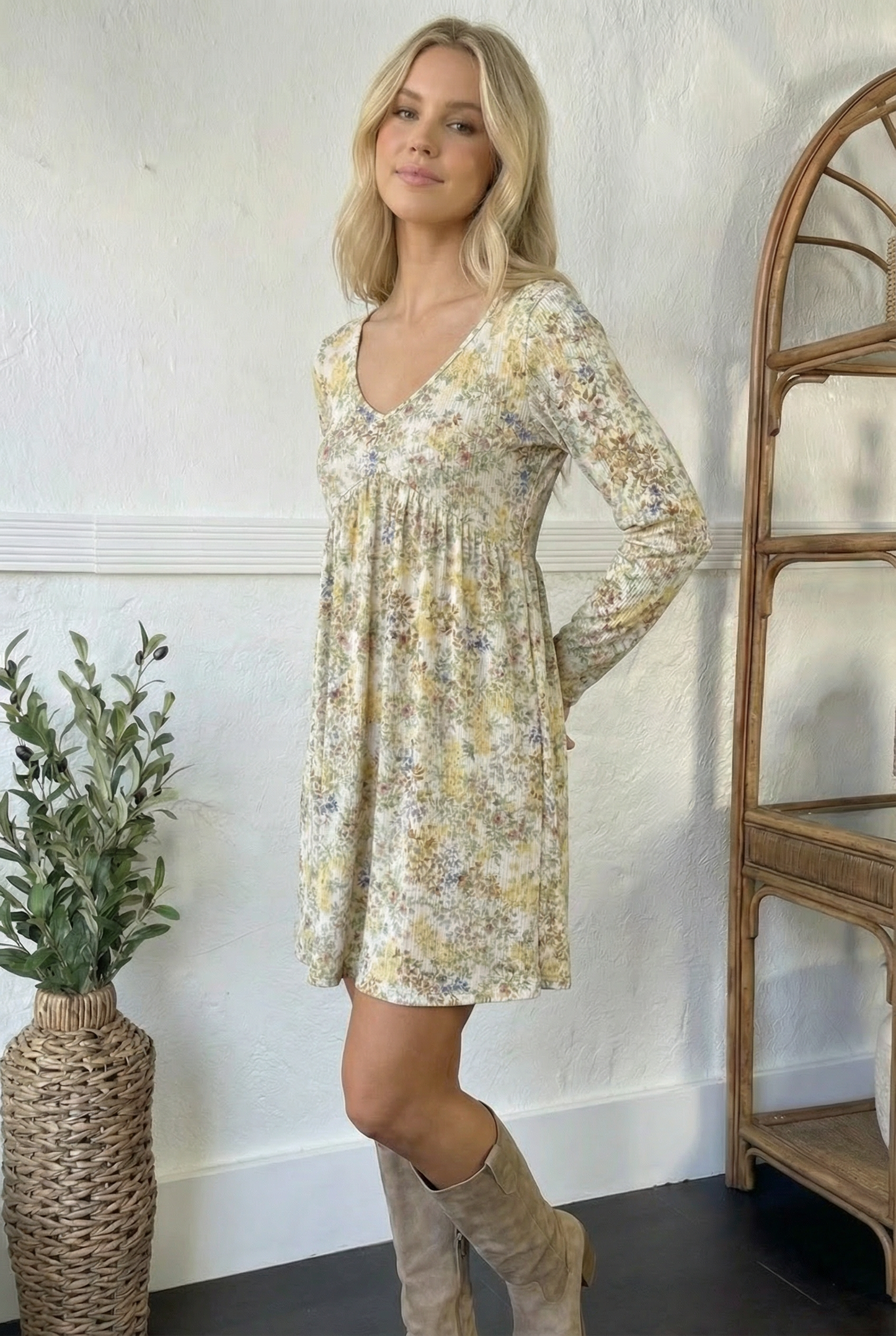 Woman wearing a floral dress standing in a room with a plant and wooden shelf.