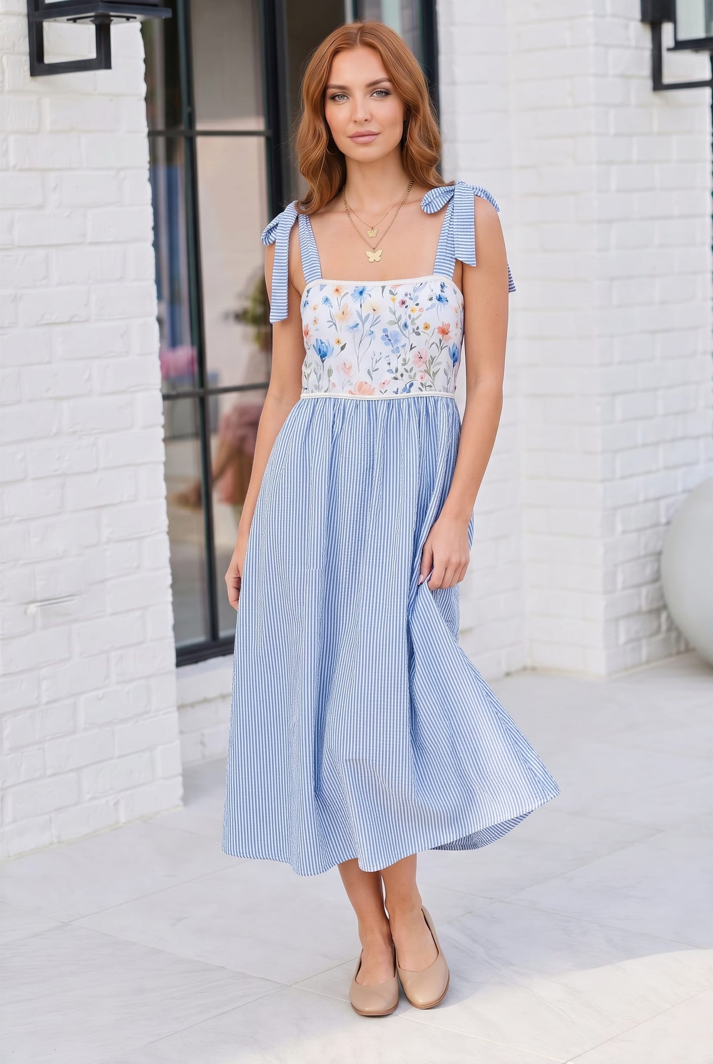 Woman wearing a blue and white dress with floral top against a white brick wall.