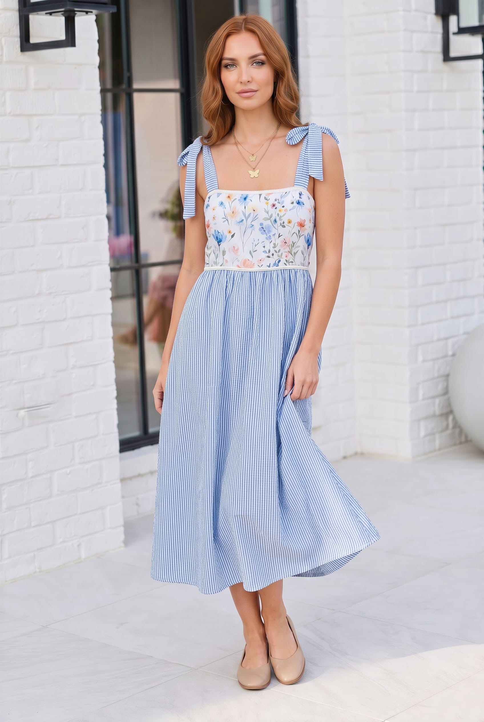Woman wearing a blue and white dress with floral top against a white brick wall.