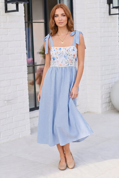 Woman wearing a blue and white dress with floral top against a white brick wall.