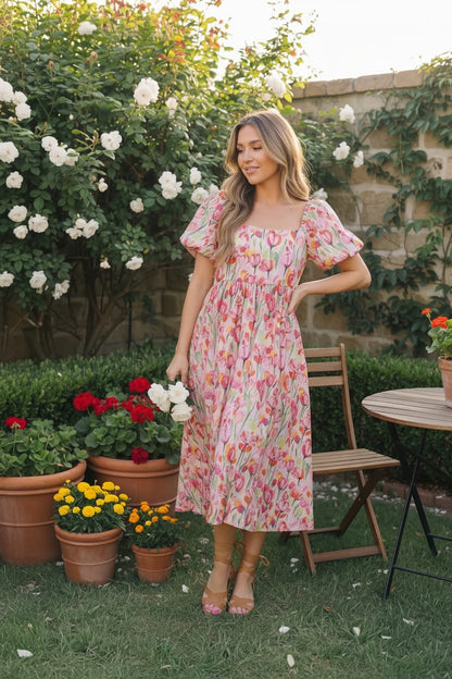 Woman in a floral dress standing in a garden with potted plants and flowers.