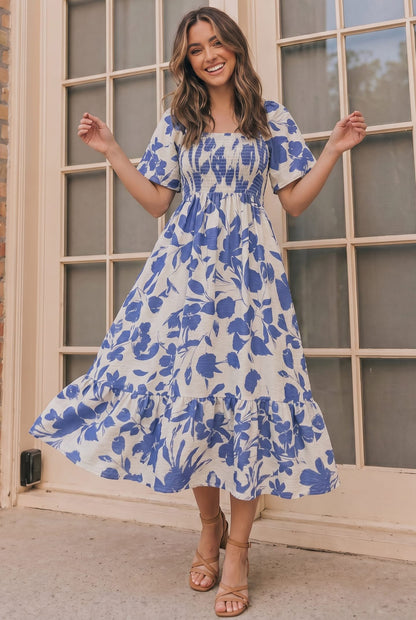 A woman wearing a blue and white floral print midi dress with puff sleeves and a smocked bodice, standing in front of a beige windowed door.