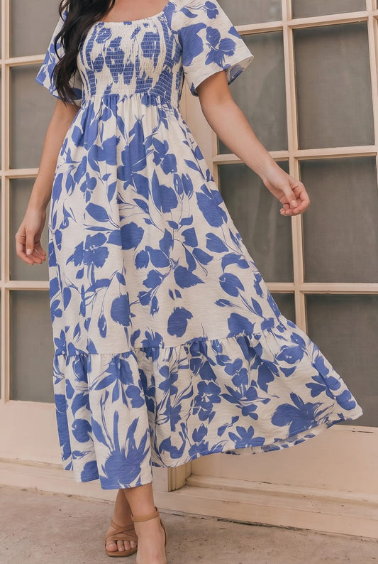 A woman wearing a blue and white floral tiered maxi dress with short flutter sleeves and a smocked bodice, standing in front of a window with wooden frames.