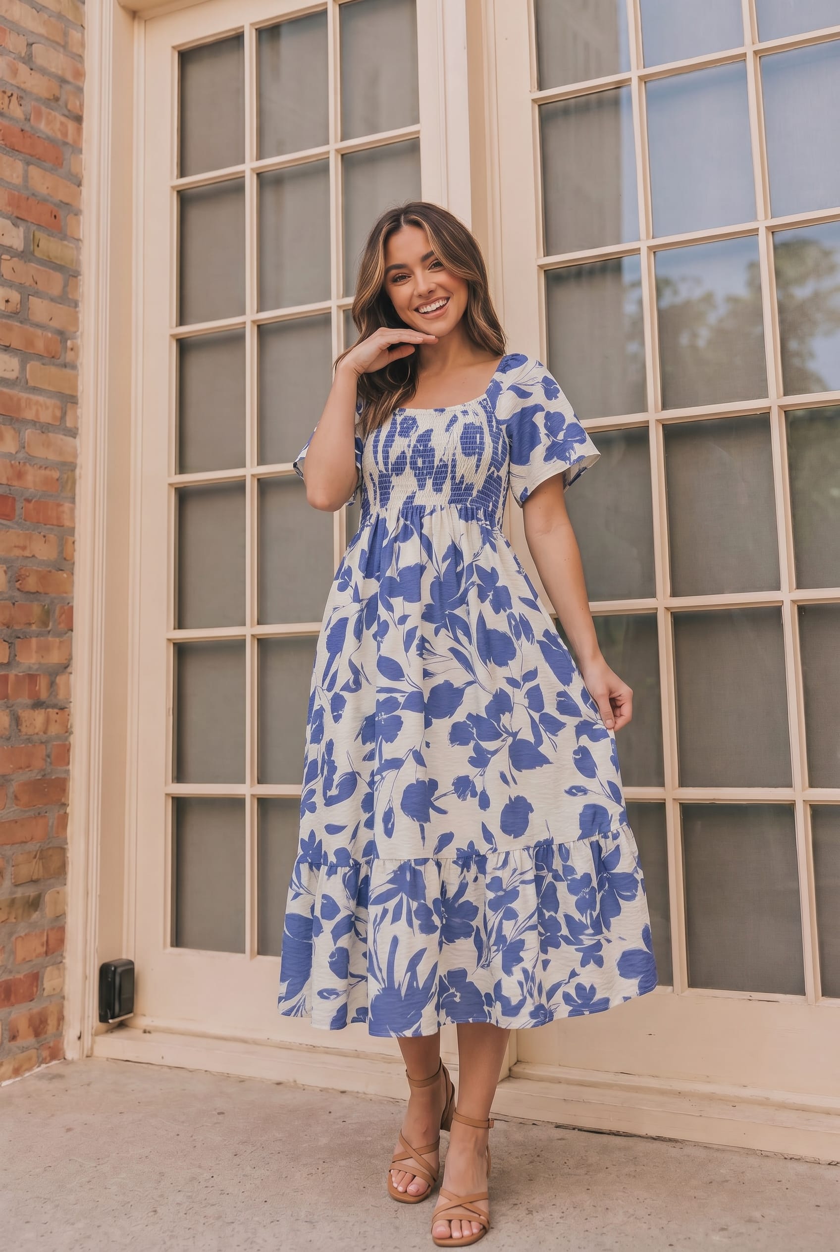 A woman wearing a blue and white floral midi dress with short puff sleeves and a smocked bodice, standing in front of a beige-framed glass door.